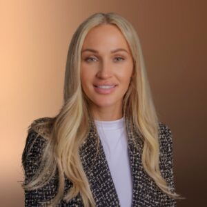 Professional headshot of a blonde woman wearing a patterned blazer and white top, smiling against a warm neutral background.