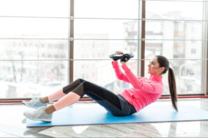 Woman performing a seated core exercise with a weight plate on a fitness mat near large windows, representing guided physical activity, strength development, and active lifestyle support during medically supervised weight loss treatment.